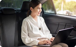 Woman in white outfit smiles while riding comfortably in the backseat of a luxury car, holding a laptop—enjoying a calm, stress-free journey.