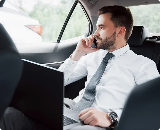 Young businessman in a white shirt and tie sits in the backseat of a car, talking on the phone while working on a laptop—portraying a focused and professional travel environment.