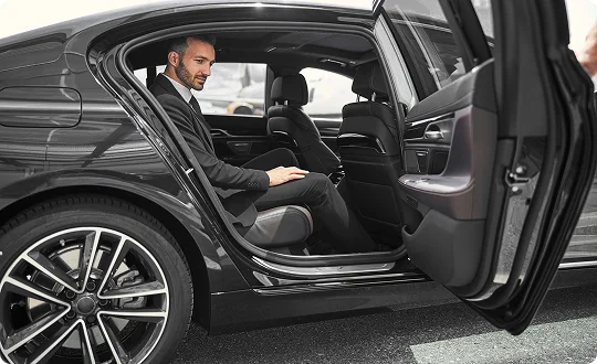 Well-dressed man settles into the backseat of a sleek black luxury sedan with the door open—capturing a smooth, professional arrival framed by polished wheels and refined interior details.