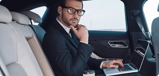 Confident businessman in a dark suit and glasses sits in the backseat of a luxury vehicle, working on a laptop with a coffee cup beside him—portraying focus, productivity, and executive comfort during transit.