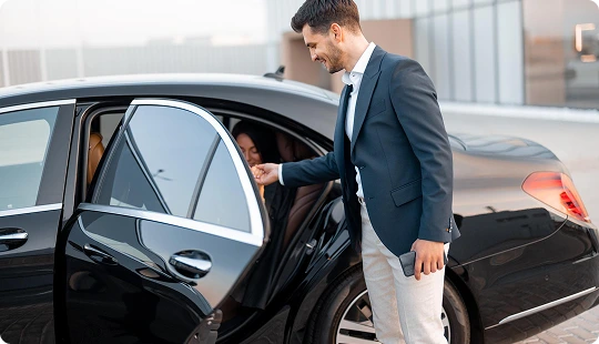 Smiling chauffeur opens the door of a luxury black car for a female passenger—reflecting the warmth, care, and professionalism of a premium chauffeur service experience.