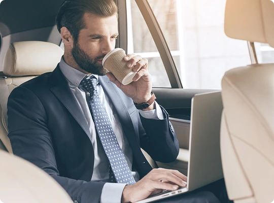 Businessman in a suit works on a laptop and sips coffee from the backseat of a luxury car—capturing a moment of comfort, focus, and productivity while on the move.