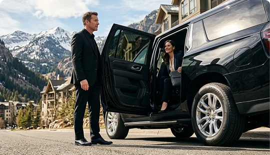 A chauffeur in a black suit holds open the rear passenger door of a black luxury SUV. A woman in a dark business suit steps out of the vehicle against a scenic backdrop of snow-dusted mountains and alpine buildings.
