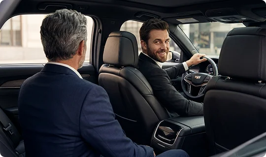 A smiling chauffeur in a suit looks back from the driver's seat of a luxury vehicle toward a passenger. The passenger sits in the backseat, seen from behind, as they converse inside the car's black leather interior.